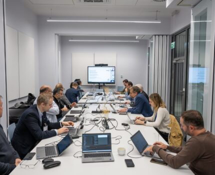 Kilkanaście osób siedzi po obu stronach długiego stołu w niedużej sali konferencyjnej. Na stole, przed uczestnikami leżą otwarte laptopy.