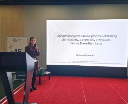 Kobieta z mikrofonem w ręku stoi na czerwonym podeście, obok na dużym ekranie widoczny slajd prezentacji z wyraźnym napisem: Optymalizacja warunków procesu ekstrakcji pierwiastków z pierników przy użyciu metody Boxa-Behnkena.