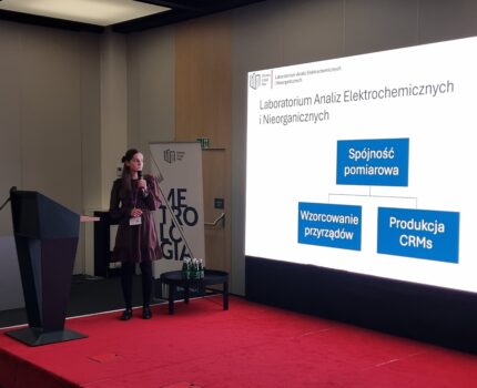 Szczupła kobieta z mikrofonem w ręku stoi na czerwonym podeście, obok na dużym ekranie widoczna prezentacja.