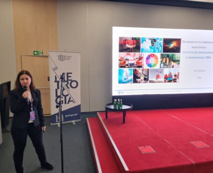 Kobieta z mikrofonem w ręku stoi obok czerwonego podestu. Przy podeście ekran z wyświetloną prezentacją.