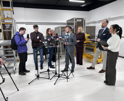 Osiem osób w laboratorium. Jeden z mężczyzn w szarym garniturze stoi przed ustawionymi mikrofonami.