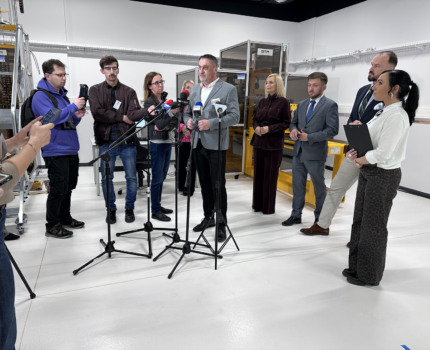 10 osób w pomieszczeniu laboratoryjnym. Mężczyzna w szarym garniturze stoi przed ustawionymi mikrofonami.