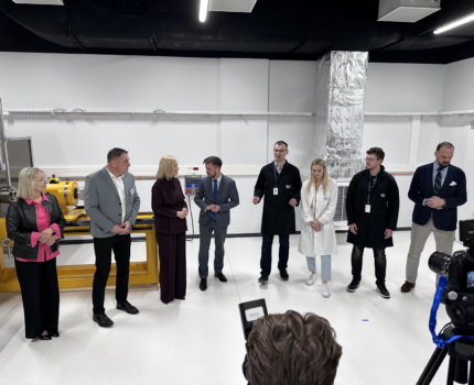 Osiem osób stoi w jednym rzędzie w pomieszczeniu laboratoryjnym. Na nich skierowana jest kamera na statywie.