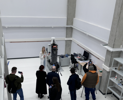 Widok z góry na wysokie, jasne pomieszczenie, w którym kobieta w białym fartuchu stoi przed grupą siedmiu osób. W dłoniach niektórych z nich widoczne telefony. Na statywie kamera.