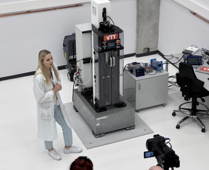 Widok z góry na wysokie, jasne pomieszczenie, w którym kobieta w białym fartuchu stoi przed kamerą ustawioną na statywie.