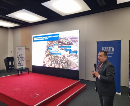 Mężczyzna z mikrofonem w ręku stoi obok czerwonego podestu. Przy podeście ekran z wyświetloną prezentacją.