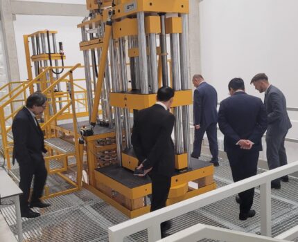 Kilku mężczyzn w ciemnych garniturach chodzi po metalowym kratkowanym podeście wokół dużego stanowiska pomiarowego wzorców masy.