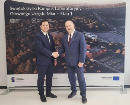 Dwóch mężczyzn w ciemnych garniturach ściska sobie ręce w geście gratulacji na tle makiety Kampusu GUM.