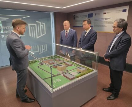 Czterech mężczyzn ubranych w ciemnoszare garnitury stoi pochylonych nad oszkloną gablotą, w której znajduje się makieta Kampusu GUM.