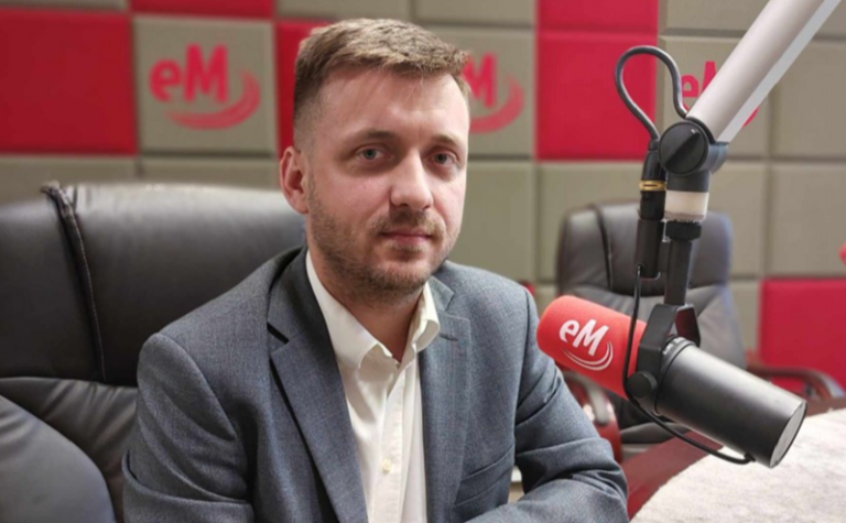 Mężczyzna w szarym garniturze w studiu radiowym. Przed nim wysunięty mikrofon z logo stacji radiowej.