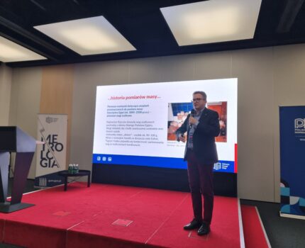 Mężczyzna w okularach, z mikrofonem w ręku stoi na czerwonym podeście, za nim duży ekran z wyświetloną prezentacją.