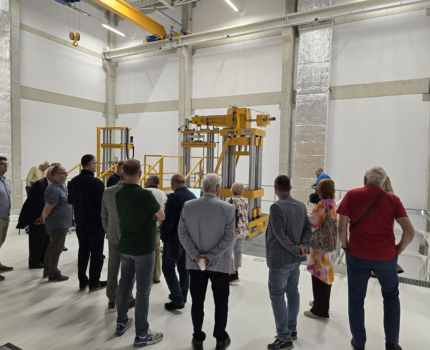 Grupa ludzi przygląda się dużym przyrządom pomiarowym stojącym w jasnym pomieszczeniu laboratoryjnym.