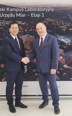 Dwóch mężczyzn w ciemnych garniturach ściska sobie ręce w geście gratulacji na tle makiety Kampusu GUM.